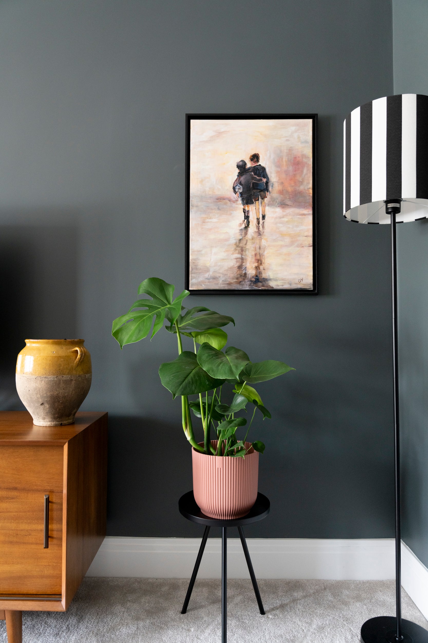 Up close image of a painting of two boys walking with a football under their arm, placed on a dark wall in a snug. There is a black and white striped lamp and a beautiful mid-century console table with a French Confit pot that is yellow.