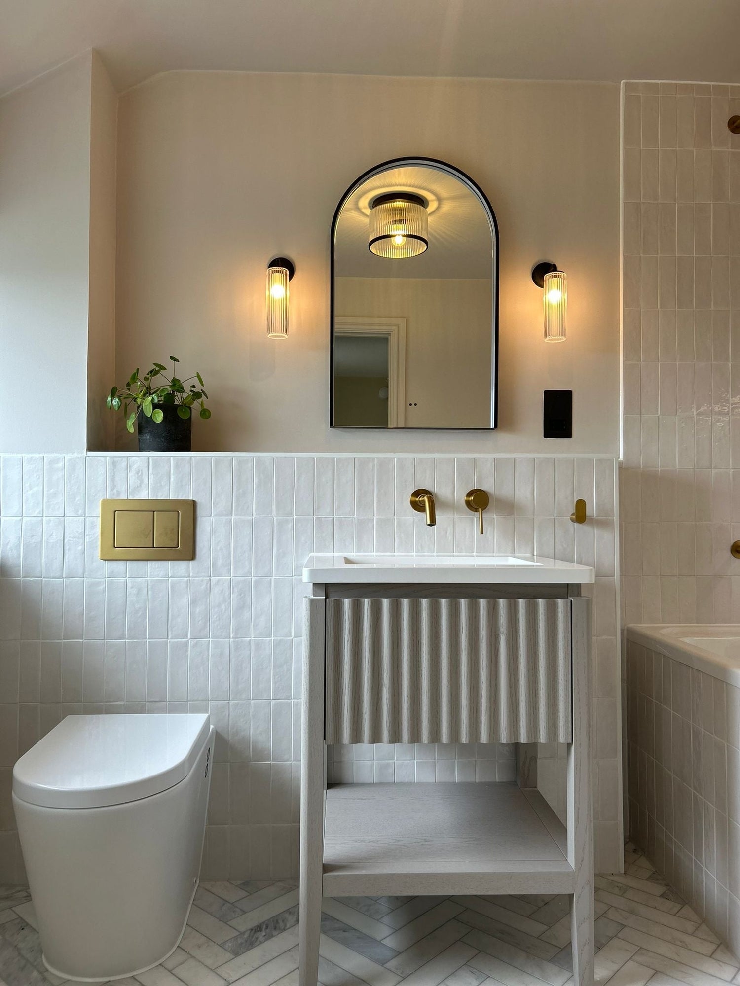 Bathroom with marble flooring, white zellige wall tiles, brass finishes, wall lighting and a fluted wooden cabinet. 