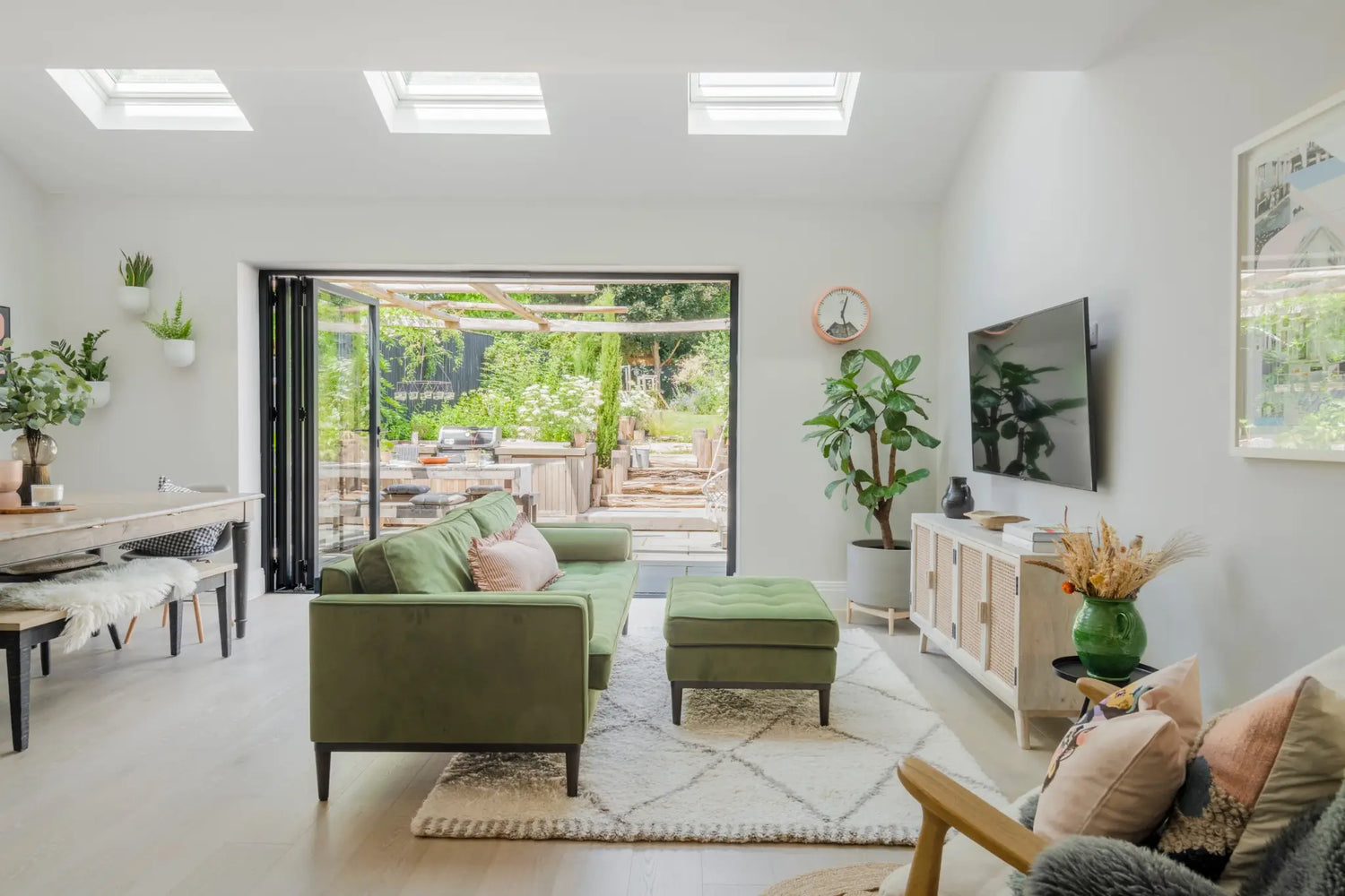 A wide angle view of an open space living/dining room with large bifolding doors that are opened onto the outdoor kitchen. There is a green velvet sofa with Soho House style decor throughout. The space is light and airy.