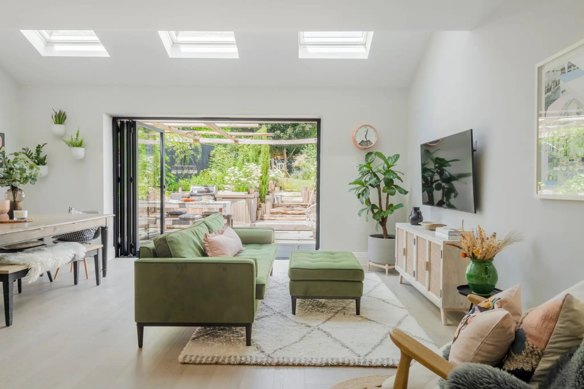 A wide angle view of an open space living/dining room with large bifolding doors that are opened onto the outdoor kitchen. There is a green velvet sofa with Soho House style decor throughout. The space is light and airy.